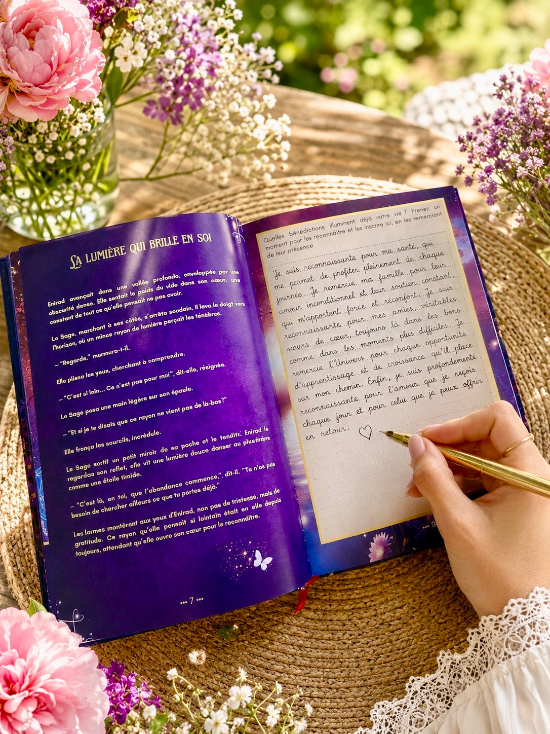 Main écrivant à la plume dorée dans le Livre Divin'Om ouvert, pages intérieures colorées visibles, entouré de roses